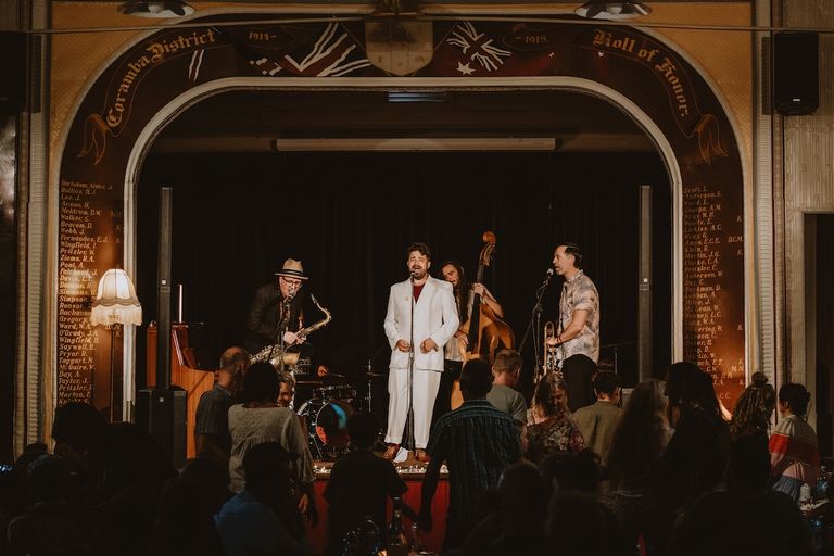 Harry James Angus sings in a white suit on stage at Coramba Hall, backed by a live band with saxophone and double bass before a dancing crowd.